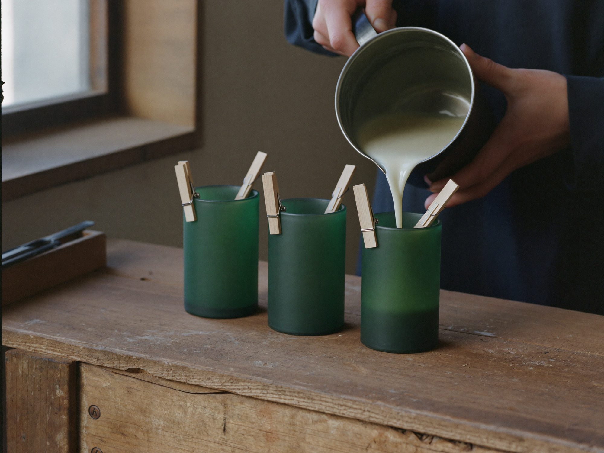 Manos vertiendo cera en vasos de vidrio verde sobre el banco del taller